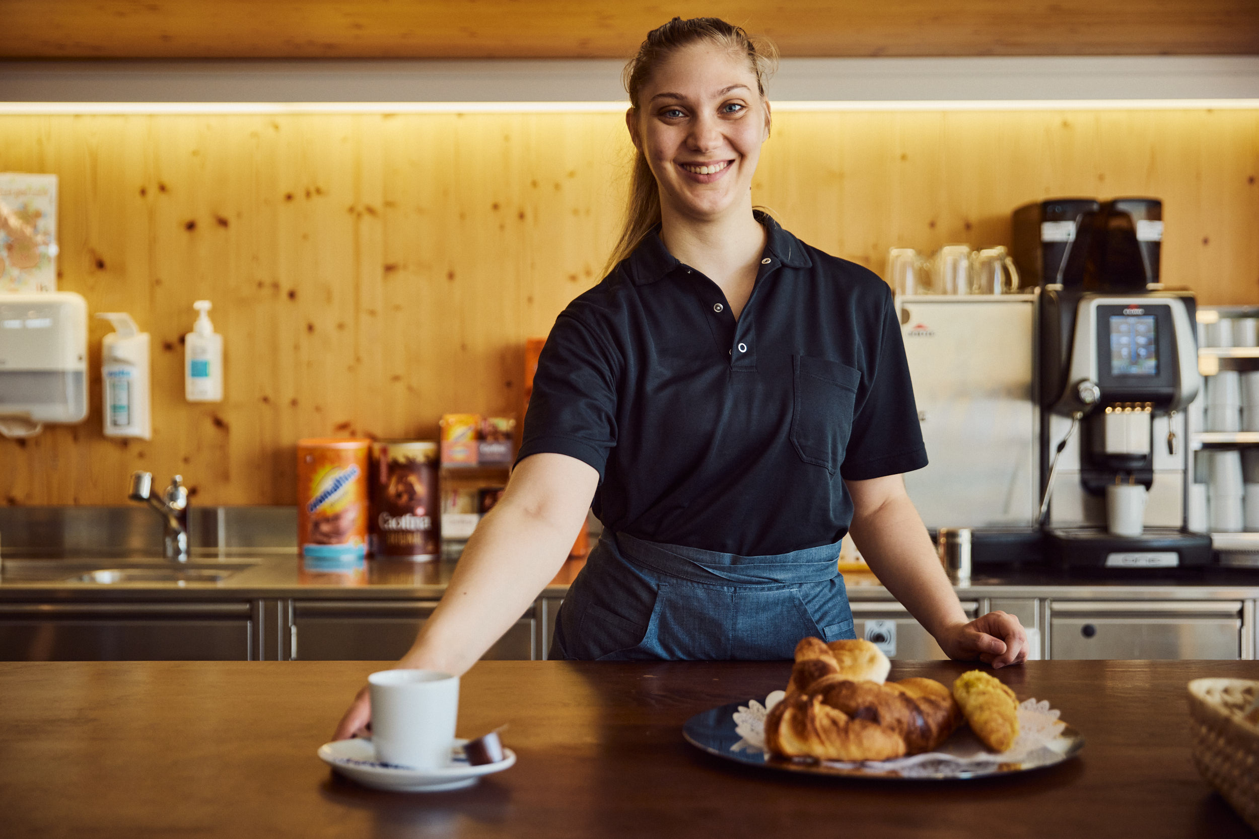 Klientin der Stiftung Lebenshilfe reicht in der Cafeteria einem Gast lächelnd einen Kaffee über die Theke – im Vordergrund frische Gipfeli.
